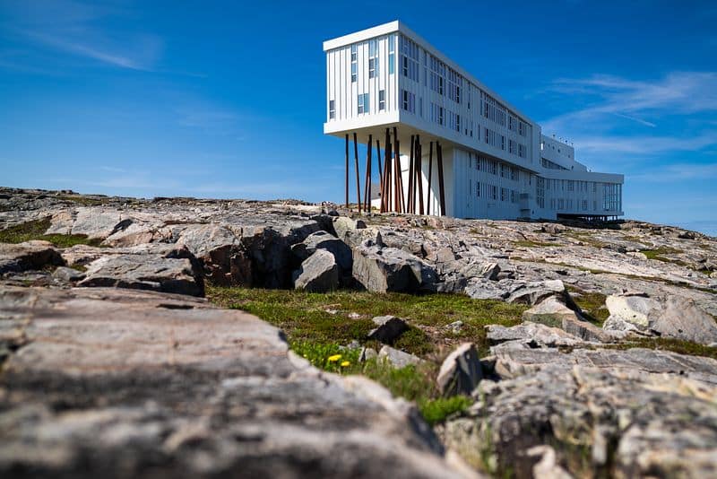 Bild von Fogo Island in Kanada: Ein modernes, weisses Gebäude mit vielen Fenstern steht auf hohen, schlanken Stelzen über einem felsigen Küstenabschnitt. Der Himmel ist klar und blau, und der Boden ist mit großen Steinen und etwas Gras bedeckt, während ein paar gelbe Blumen im Vordergrund sichtbar sind.
