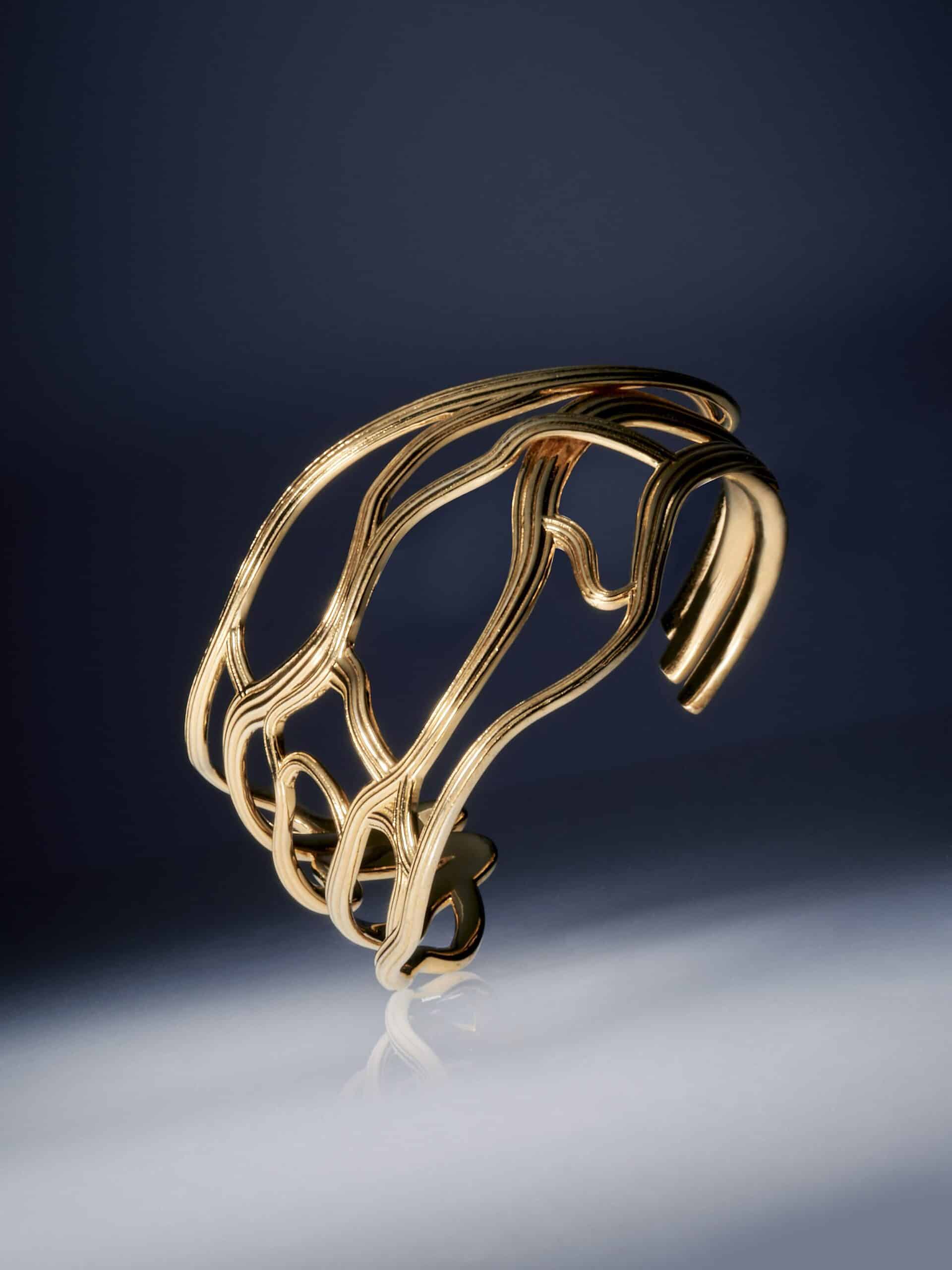 Yan Jiangs „Fine Edition“ Armreif fasziniert mit seiner kunstvoll geschwungenen Goldform, die sich elegant auf der spiegelnden Oberfläche widerspiegelt.