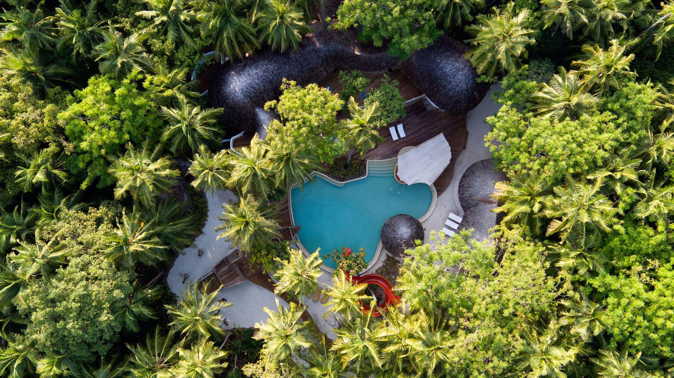 Aus der Vogelperspektive offenbart sich eine luxuriöse Poollandschaft mit roter Wasserrutsche, eingebettet in ein Meer aus sattgrünen Palmen und tropischer Vegetation, die zum Verweilen einlädt.