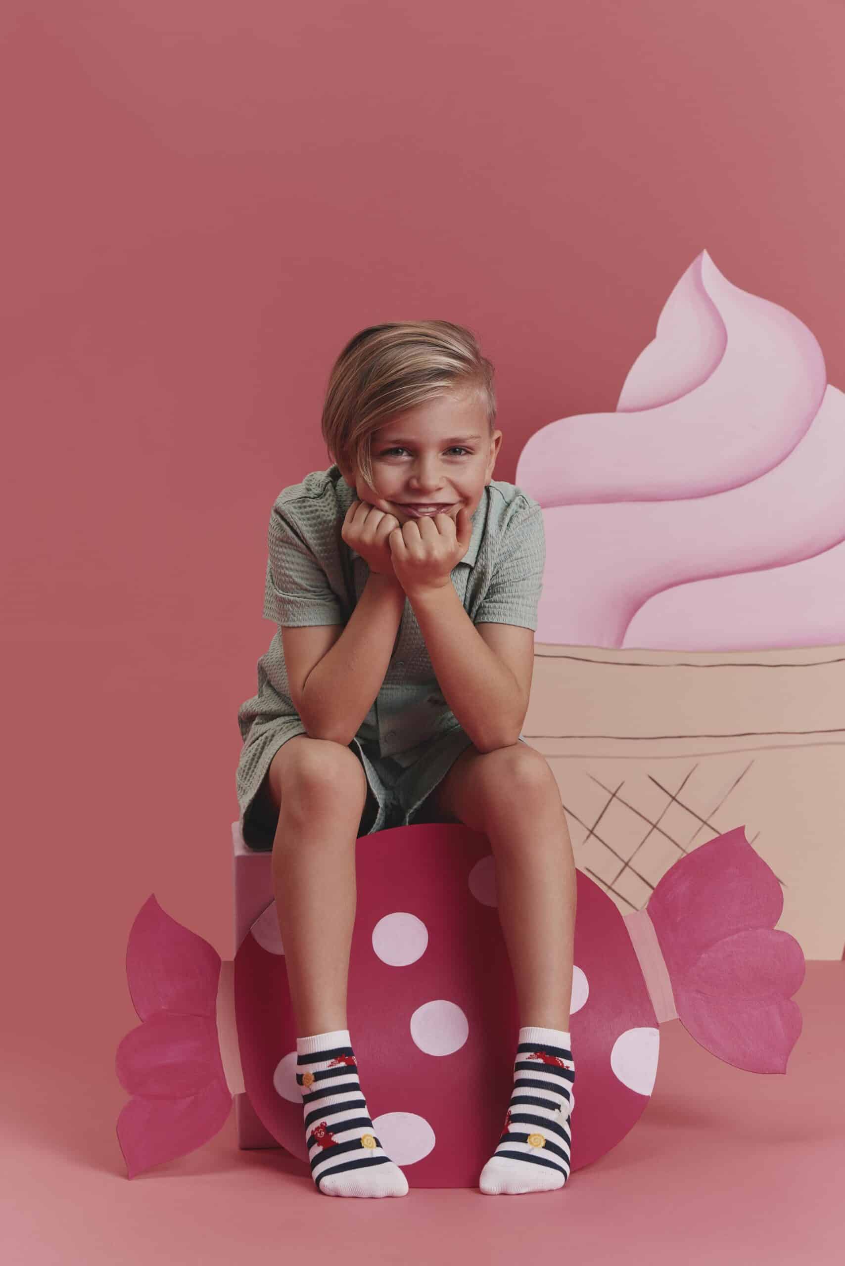 Fröhliche FALKE Legwear für SS26: Ein Kind mit gestreiften Socken und einem bezaubernden Lächeln posiert spielerisch zwischen bunten Sussigkeiten und Eis.