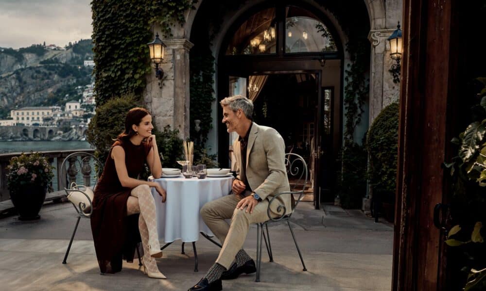 Ein elegantes Paar genieSSt einen lebhaften Austausch auf einer idyllischen Terrasse, wahrend ihre anspruchsvollen Strumpfe und Socken den stilvollen FALKE SS26-Look perfekt erganzen.