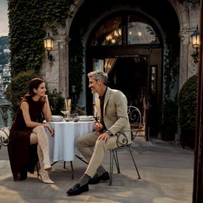 Ein elegantes Paar genieSSt einen lebhaften Austausch auf einer idyllischen Terrasse, wahrend ihre anspruchsvollen Strumpfe und Socken den stilvollen FALKE SS26-Look perfekt erganzen.