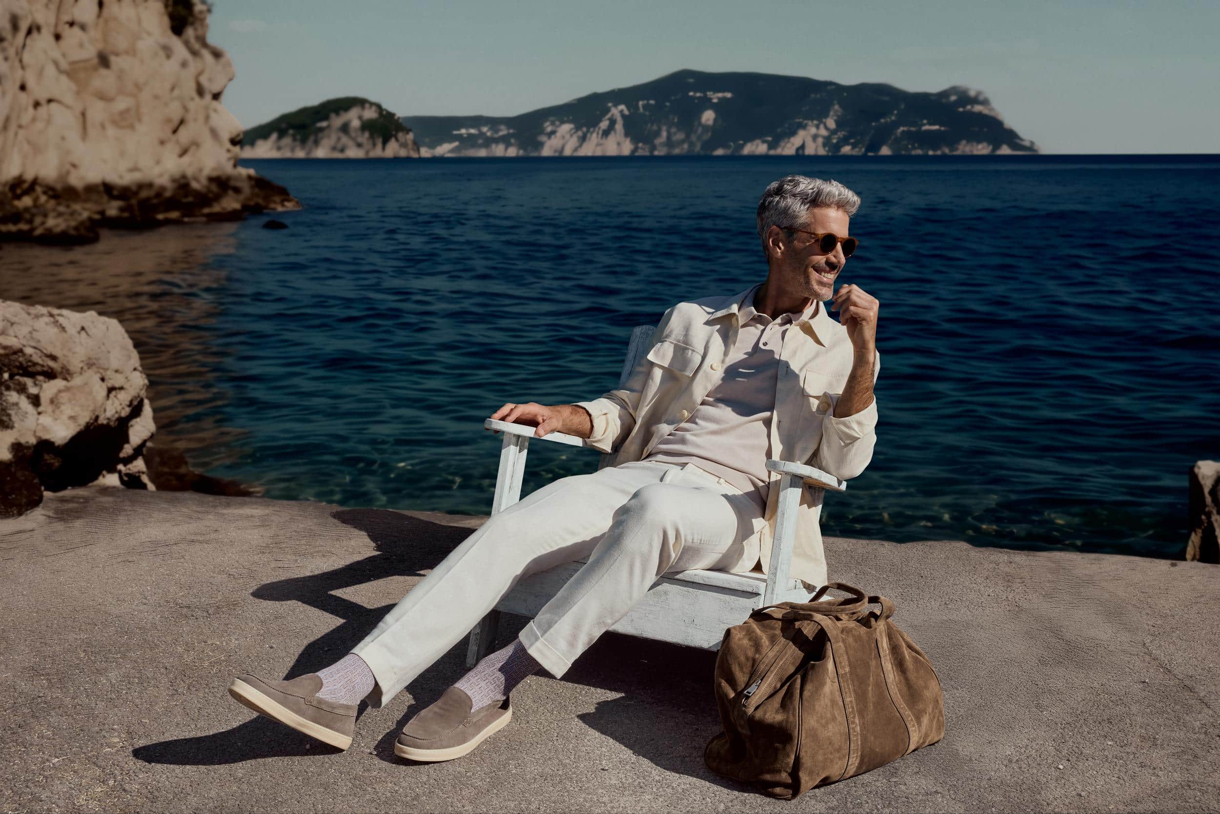 In leichter Kleidung und mit dezent gemusterten FALKE Socken entspannt ein Mann lachend an einem sonnigen Meerblick, passend zur SS26 Kollektion.