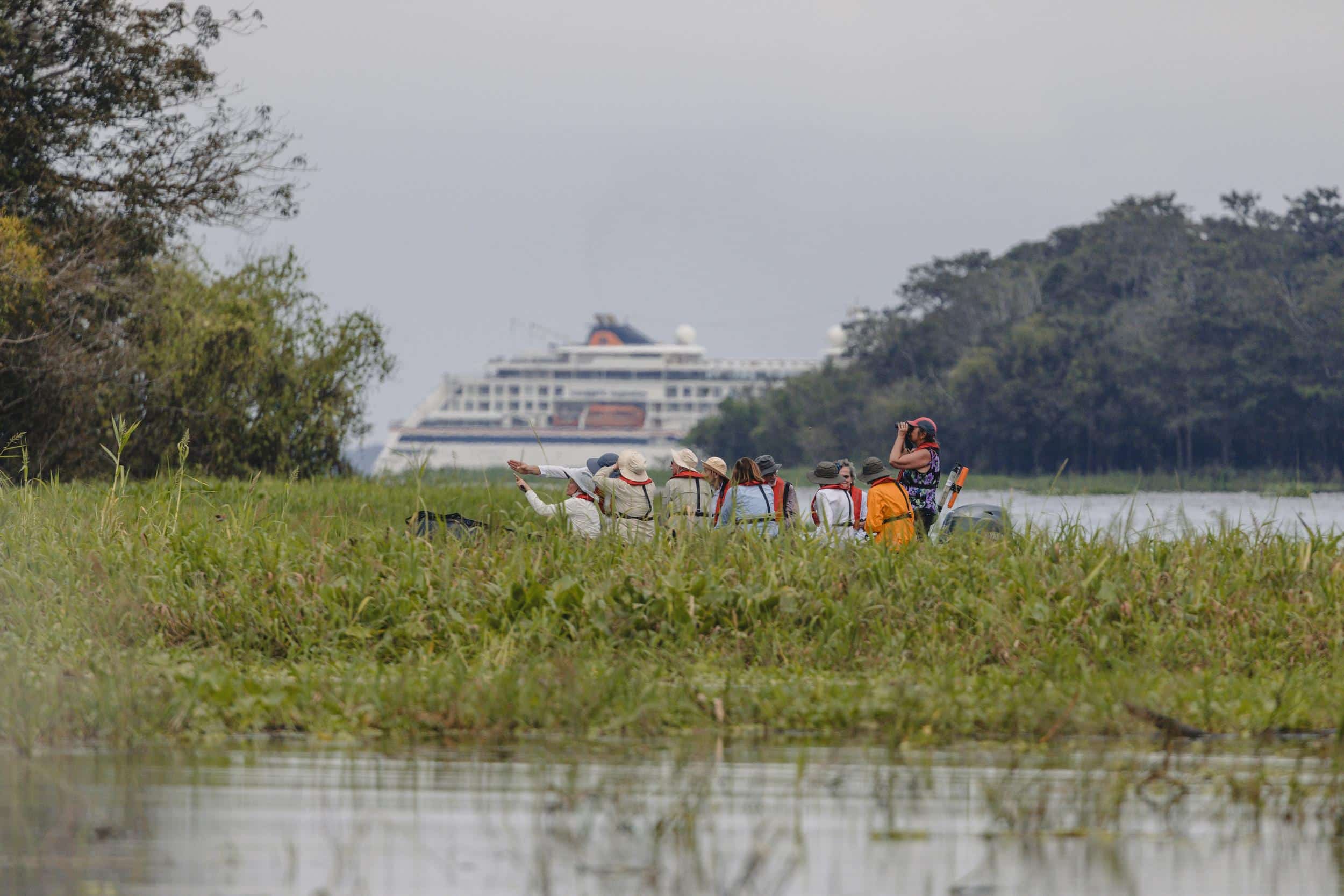 Auf einer Hapag-Lloyd Expedition am Amazonas blicken Reisende vom Beiboot aus auf die beeindruckende Flusslandschaft, im Kontrast zum majestätischen Kreuzfahrtschiff am Horizont.